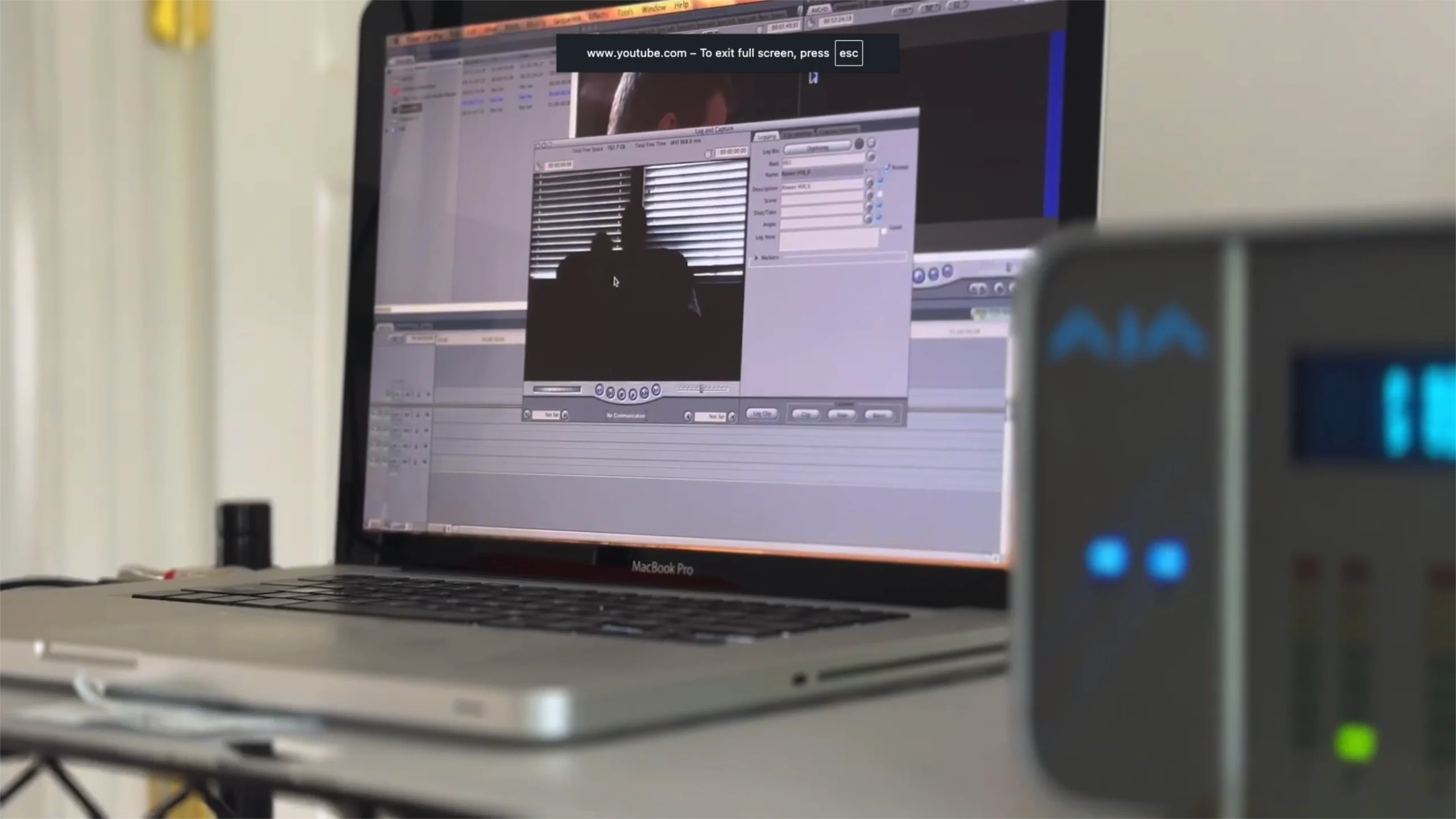 MacBook Pro open to video editing software with timeline and multiple panels on screen, plus a gray external device with blue LEDs on the right.